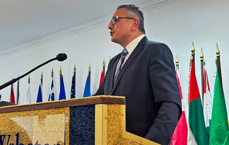 A man speaks at a Webster University podium with several country flags behind him.