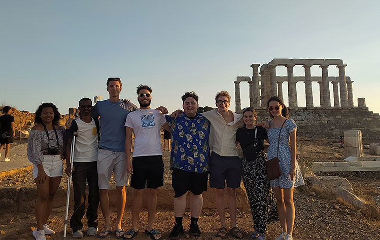 Students studying at Webster Athens stand in front of the ruins of the Temple of Poseidon at sunset.