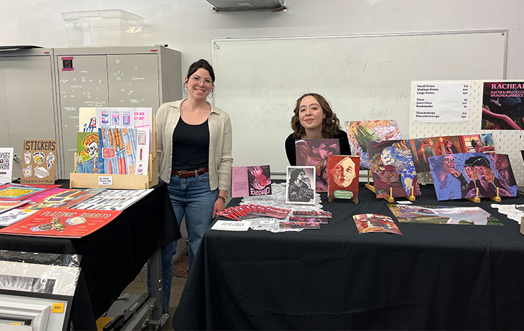 Two women smile for a photo while posing with their art booths.