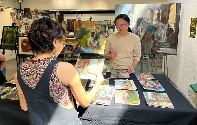 An artist stands behind a table while an attendee holds a painting from the booth.