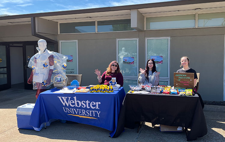 Three people sit at a Webster branded table on a sunny day outside the Webster University Visual Arts Building.