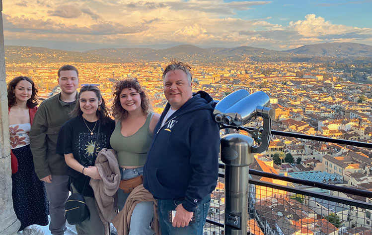 A group from Webster smiles for a photo while standing next to a pair of binoculars on the roof of the Duomo, overlooking the landscape of Florence.