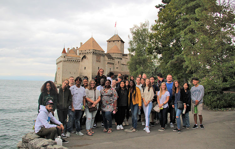 A group of Webster students stand in front of the Chateau De Chillon on a cloudy day.