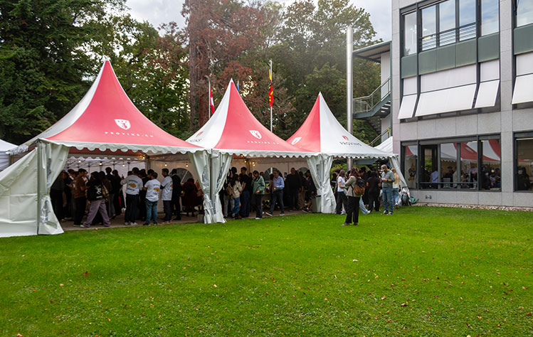 Three red and white tents sit along the sidewalk next to grassy patch outside of a Webster Geneva Campus building.