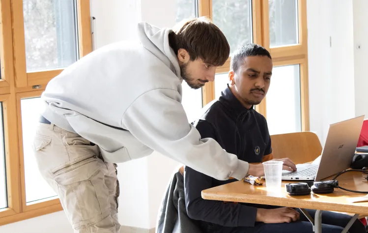 Two teammates look at a laptop in a room with windows directly behind them.