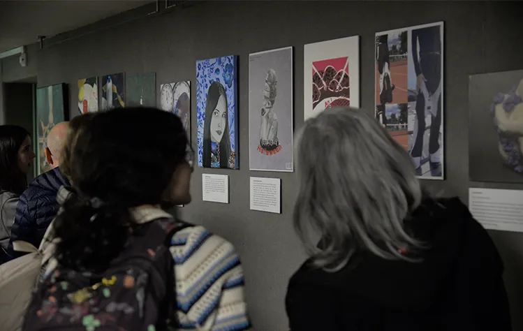 Two attendees view student artwork along a grey wall in a dark room.