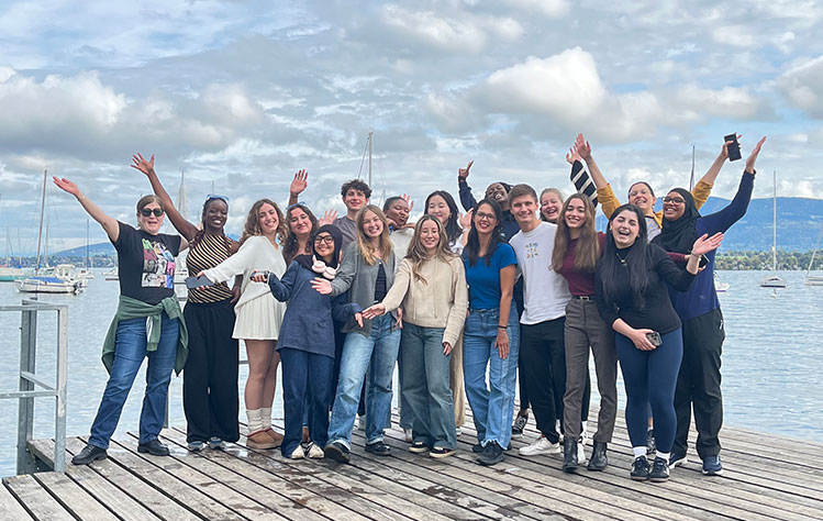 Smiling students stand on a dock in front of a lake with a view of mountains in the background.