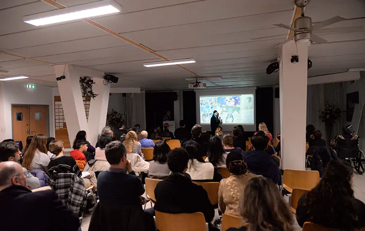 Media showcase attendees sit in a room facing a projector where a student presents.