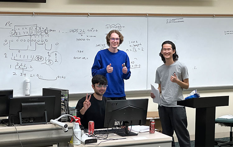 The Golden Gorloks team surrounds a computer to solve a problem. Math equations are written on the whiteboard behind them.