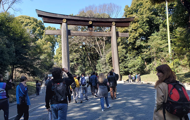 A group of people walk under a large torii gate on a pathway in a wooded area in Japan.