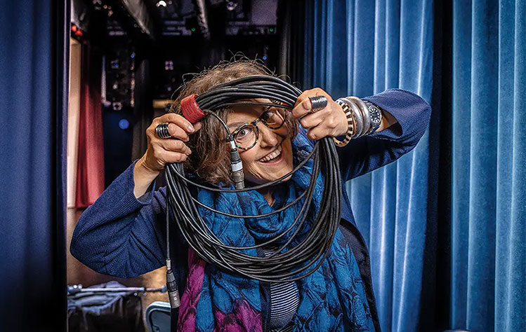 Photo of Joan Lipkin holding wires for a microphone behind the curtains on a stage.