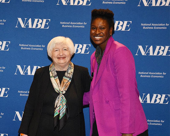 Kristen McLauchlin, dressed formally, poses in front of a NABE step and repeat with another person.