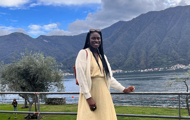 Kristen McLauchlin wears a yellow dress while smiling in front of a body of water and mountains with greenery.