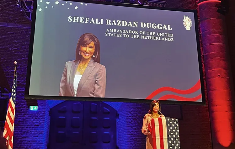 Shefali Razdan Duggal stands onstage at a podium to speak with a screen displaying her name and photo behind her.