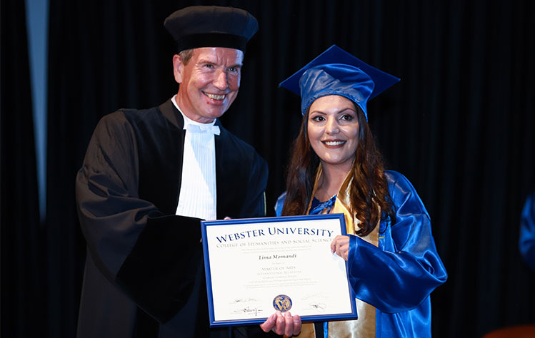 Rector Jean-Paul Marissing and Lima Momandi hold Momandi's diploma onstage together while wearing formal commencement dress.