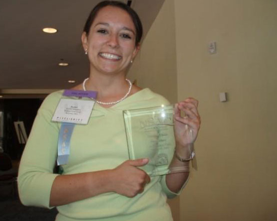 McInnisi holds a glass plaque that reads NSAC while wearing a nametag and competition ribbon.