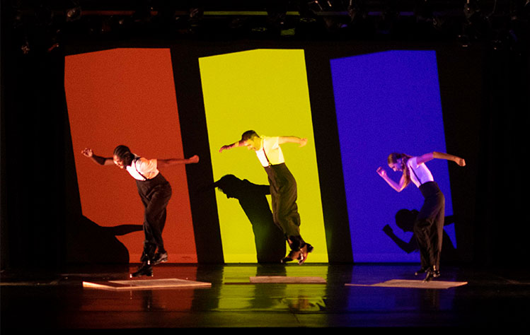Three dancers - each in front of a different colored backdrop. The backdrop colors are red, yellow, and blue.