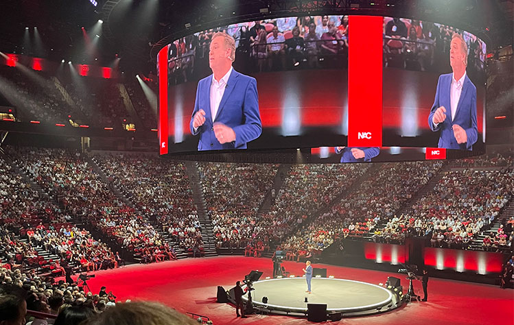 A man stands on a stage in a large arena. His image is displayed on a large circular screen at the top of the arena.