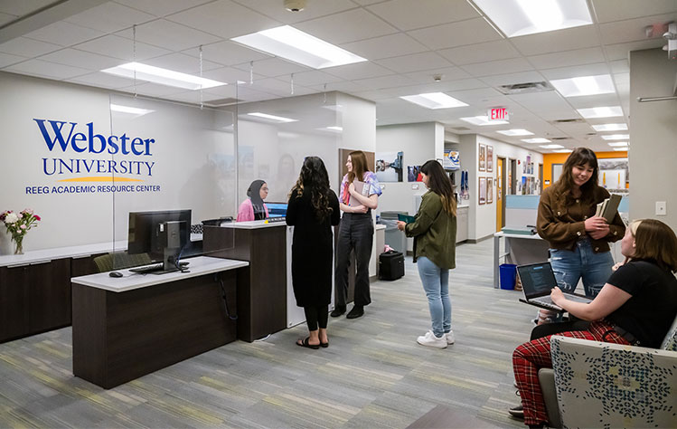 Students sit in chairs around the room and stand near the Reeg Academic Resource Center's welcome desk while holding books and laptops.