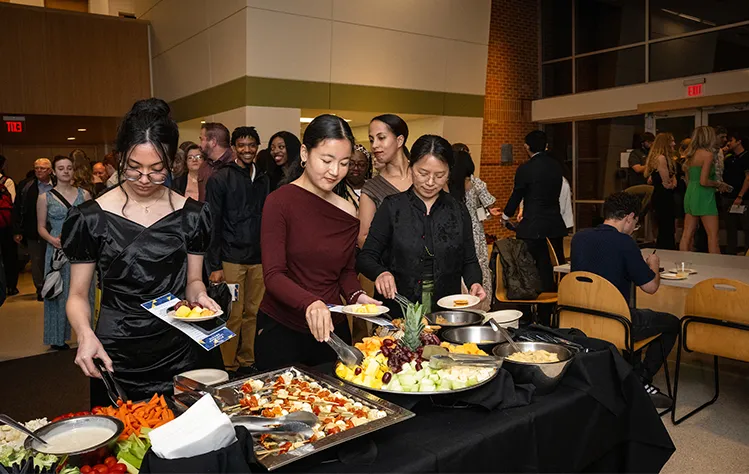 Three students fill their plates with food from the buffet line in the atrium of the Community Music School building on the Webster Groves campus.