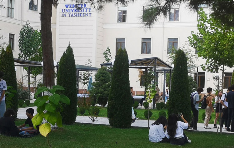 Webster Tashkent students congregate in a grassy common area of campus with the Webster Tashkent building and sign in the background.