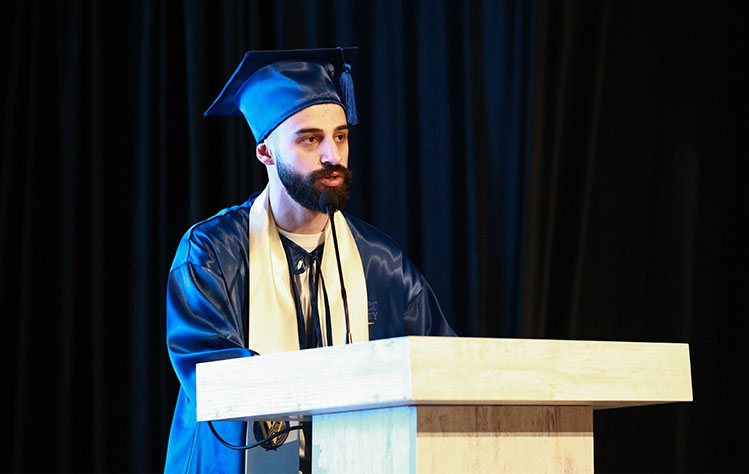  Vano Gazdeliani stands at a podium to speak while wearing a Webster blue graduation cap, gown and regalia..