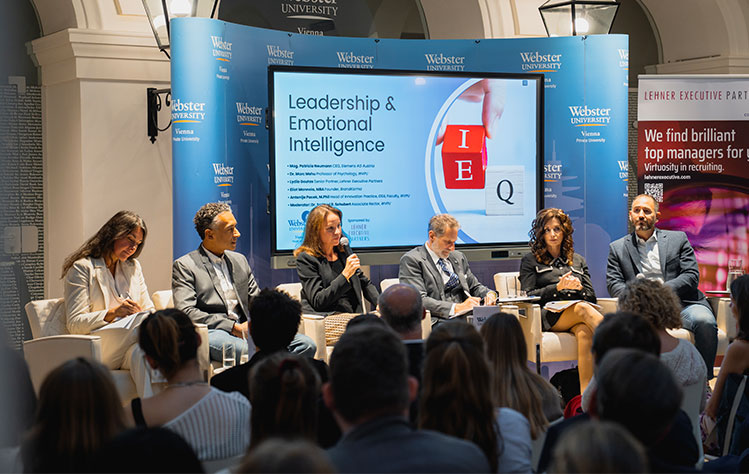 Panelists sit onstage in front of a Powerpoint presentation in Webster Vienna's Palais Wenkheim atrium.