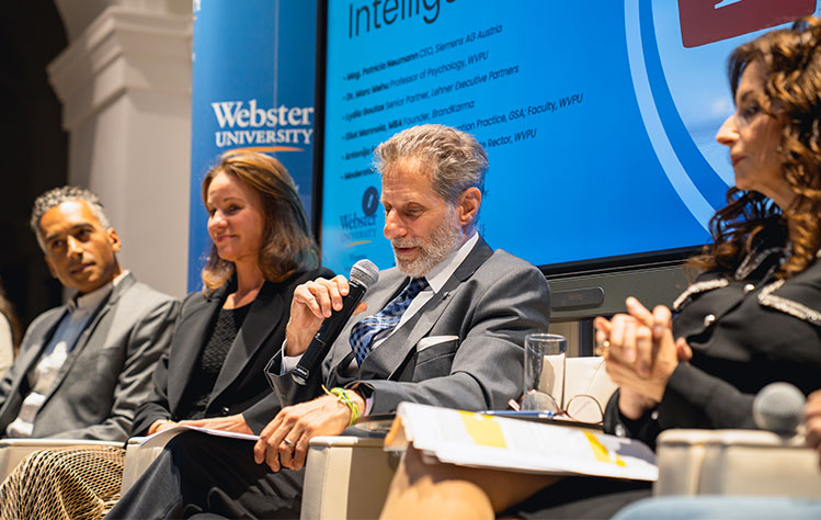 A panel member answers a question while holding a microphone. Two other panel members look on.