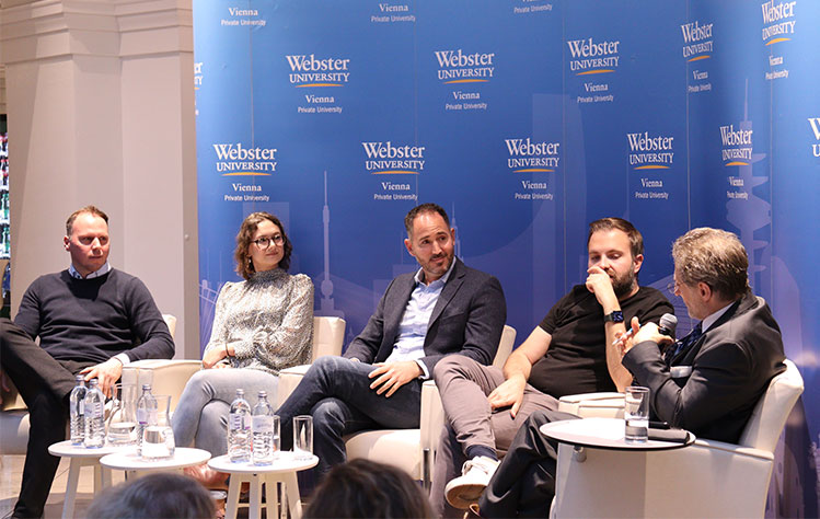 Event panelists sit in front of a blue Webster University step-and-repeat as the event begins.