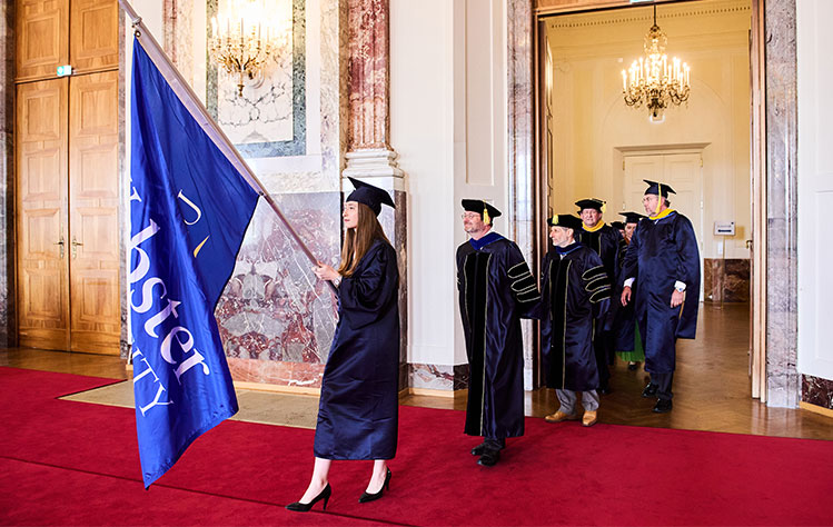 University leadership follows a graduate who leads the procession holding a Webster University flag.